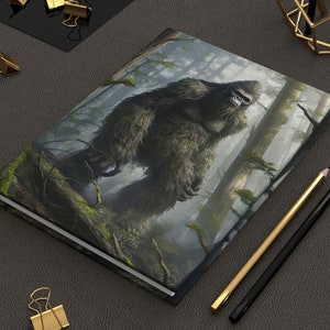 May include: A hardbound journal featuring a detailed illustration of a Bigfoot in a forest setting. The cover art depicts the creature standing among trees, with a green and brown color palette. Gold binder clips and pencils are also visible.