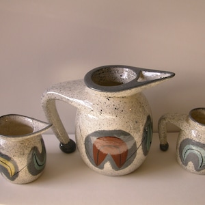 May include: A set of three ceramic jugs with a speckled white glaze and abstract geometric designs in shades of grey, brown, green, and yellow.