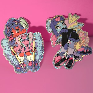 Puede incluir: Dos pegatinas holográficas con personajes de dibujos animados. La primera pegatina presenta un personaje rosa y azul con una cara roja y cuernos, sosteniendo dos palos amarillos. La segunda pegatina presenta un personaje azul con un corazón rosa en la cabeza, vistiendo un atuendo negro y azul y patines. El texto "Sugar Skull" es visible en la primera pegatina.