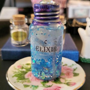 May include: A small glass bottle with a blue and purple iridescent lid filled with blue glitter and a clear liquid. The bottle has a white label that says "ELIXIR". The bottle is sitting on a white saucer with a floral design.