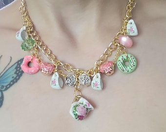 Può includere: Una collana a catena dorata con più ciondoli, tra cui tazze da tè, ciambelle e fiori. I ciondoli sono in porcellana e hanno un'estetica vintage.
