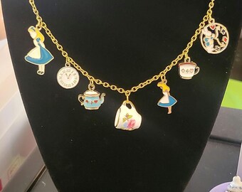 Può includere: Una collana a catena dorata con più ciondoli, tra cui personaggi di Alice nel Paese delle Meraviglie, una teiera, una tazza da tè, un orologio e una carta da gioco.