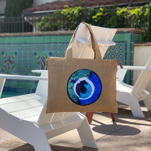 Puede incluir: Una bolsa de mano marrón con un diseño de ojo turco azul y verde. La bolsa está hecha de yute y tiene una sola correa.