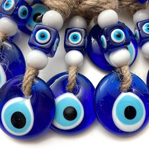 May include: A collection of blue evil eye charms with white and blue details. The charms are strung on twine and have white beads between them. The charms are in the shape of a circle and a square.