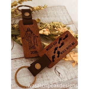 May include: Two wooden keyrings with brown leather straps and metal rings. One keyring features an engraved image of a figure and the number 2026. The other keyring has the text "Moses 624" and "Walk With Me" with footprints.