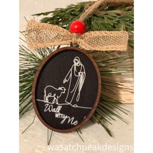 May include: Oval wooden Christmas ornament with a black background and a white line drawing of a figure with a sheep. The text "Walk with Me" is below the drawing. The ornament is decorated with a burlap bow and a red bead.
