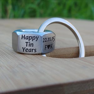 Könnte beinhalten: Ein silberfarbener Metall-Schlüsselanhänger in Form einer Mutter. Der Schlüsselanhänger trägt die Gravur "Happy Tin Years", das Datum "22.11.15" und die Initialen "F♥K". Er ist an einem silbernen Ring befestigt.