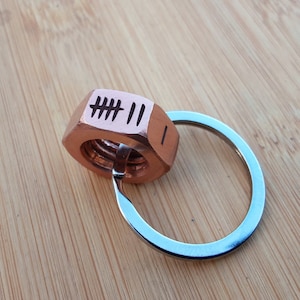 May include: A copper-coloured metal nut with the engraved text "### II" attached to a silver key ring.