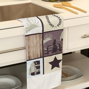 May include: A white kitchen towel with a patchwork design featuring a farmhouse theme. The design includes a barn, a star, a wreath, and a wooden plank pattern.