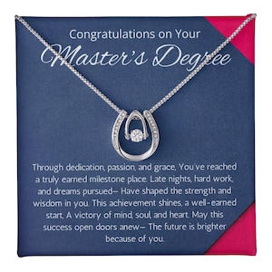 May include: A silver horseshoe pendant necklace with a central clear stone, displayed on a blue card with the text "Congratulations on Your Master's Degree". The card has a pink border and inspirational text.