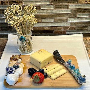 May include: A wooden cheese board with a variety of cheeses, fruits, and decorative sea-themed accents. The board features a small knife with a blue and white beaded handle.