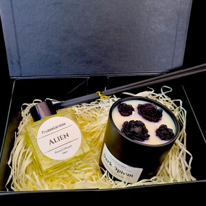 Op de afbeelding: Een zwarte cadeaudoos met een zwarte kaars en een glazen fles met rietdiffuser. De kaars heeft zwarte was met zwarte bloemaccenten en de tekst "Dark Opium". De rietdiffuserfles heeft een gele vloeistof en de tekst "Alien" en "Rietdiffuser".