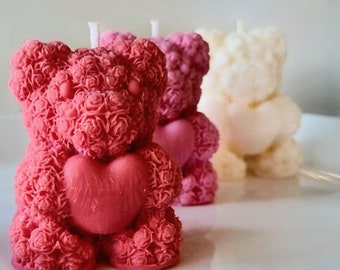 May include: Three teddy bear-shaped candles in shades of red and cream. The front candle is a deep red, with a heart-shaped detail and a textured rose pattern. The other two candles are pink and cream, with a heart detail. The candles are on a white surface.