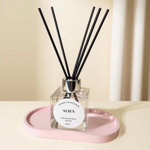 May include: A clear square glass reed diffuser bottle with a silver collar and five black reeds. The white circular label reads London Home Scents ALIEN Handmade Reed Diffuser 100ml. The bottle sits on a smooth, oval-shaped light pink tray.