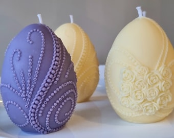 Könnte beinhalten: Zwei dekorative Osterkerzen in Eiform, eine violette mit Perlenmuster und eine weiße mit Blumenmuster.