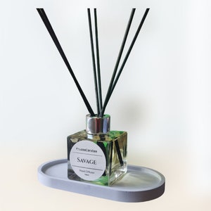May include: A clear glass bottle with a silver lid and black reeds, filled with a light yellow liquid. The bottle has a label that says "FruzzaCandles" and "SAVAGE Reed Diffuser 100ml". The bottle is sitting on a grey oval-shaped tray.