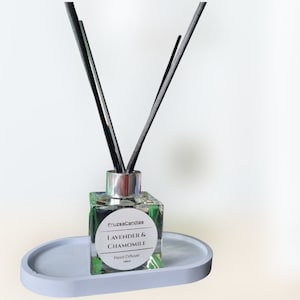 Op de afbeelding: Een heldere glazen fles met een zilveren dop en zwarte rietjes, gevuld met een lavendel- en kamille-rietdiffuser. De fles heeft een wit etiket met de tekst "FruzsasCandles Lavender & Chamomile Reed Diffuser 100ml".