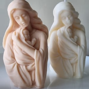 May include: Two candle sculptures of a woman holding a baby. The candle on the left is a light brown color, while the candle on the right is white.