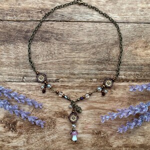 May include: A bronze-coloured necklace with a mushroom pendant and floral details. The necklace has a fine chain and is decorated with small beads in shades of purple, clear, and brown. The mushroom pendant has a white and purple cap.