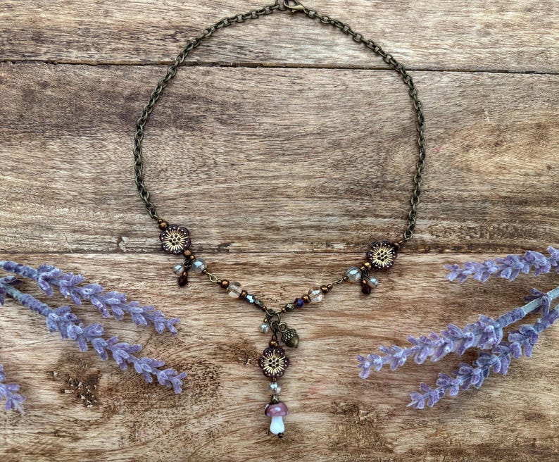 May include: A bronze-coloured necklace with a mushroom pendant and floral details. The necklace has a fine chain and is decorated with small beads in shades of purple, clear, and brown. The mushroom pendant has a white and purple cap.