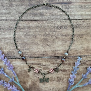 May include: A delicate bronze necklace with a dragonfly charm, accented with small flower and star charms. The necklace features brown and clear beads.