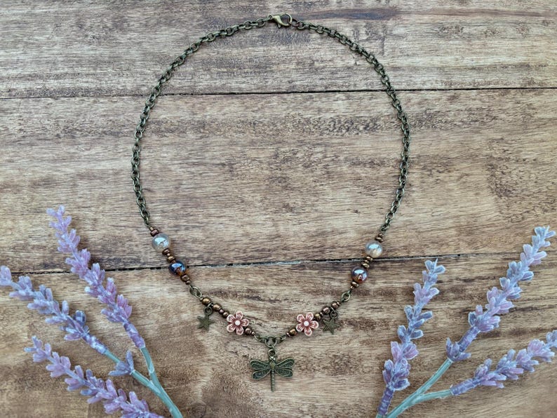 May include: A delicate bronze necklace with a dragonfly charm, accented with small flower and star charms. The necklace features brown and clear beads.