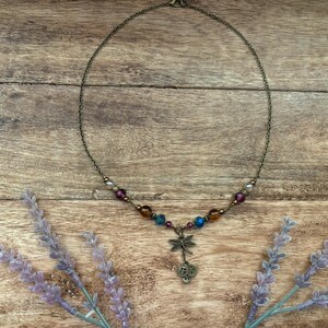 Puede incluir: Un delicado collar con una cadena de bronce y un colgante de lib&eacute;lula. El collar presenta cuentas coloridas en tonos morado, azul y marr&oacute;n. El fondo es una superficie de madera con ramitas de lavanda.