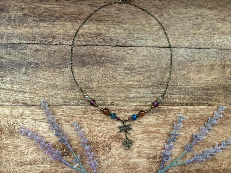 Puede incluir: Un delicado collar con una cadena de bronce y un colgante de lib&eacute;lula. El collar presenta cuentas coloridas en tonos morado, azul y marr&oacute;n. El fondo es una superficie de madera con ramitas de lavanda.