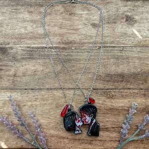 May include: Two silver necklaces with matching pendants. Each pendant depicts a couple kissing, with red and white accents. A red teardrop-shaped bead hangs from each necklace. The necklaces rest on a wooden surface.