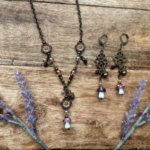 May include: A bronze-toned necklace and earrings set with a nature-inspired design. The necklace and earrings feature floral accents and mushroom charms. The earrings have an openwork design with acorn and mushroom pendants. The necklace has a similar design with a mushroom pendant.