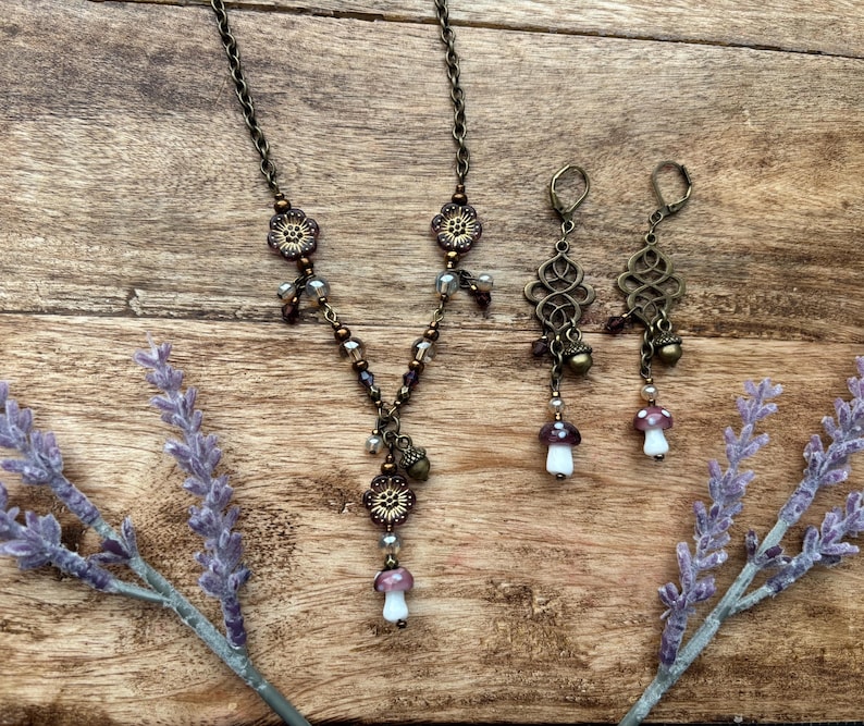 May include: A bronze-toned necklace and earrings set with a nature-inspired design. The necklace and earrings feature floral accents and mushroom charms. The earrings have an openwork design with acorn and mushroom pendants. The necklace has a similar design with a mushroom pendant.