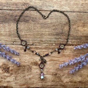 May include: A bronze necklace with a heart-shaped chain. It features floral and mushroom pendants, plus small beads in purple, brown, and clear. Lavender sprigs are placed on a wooden surface.