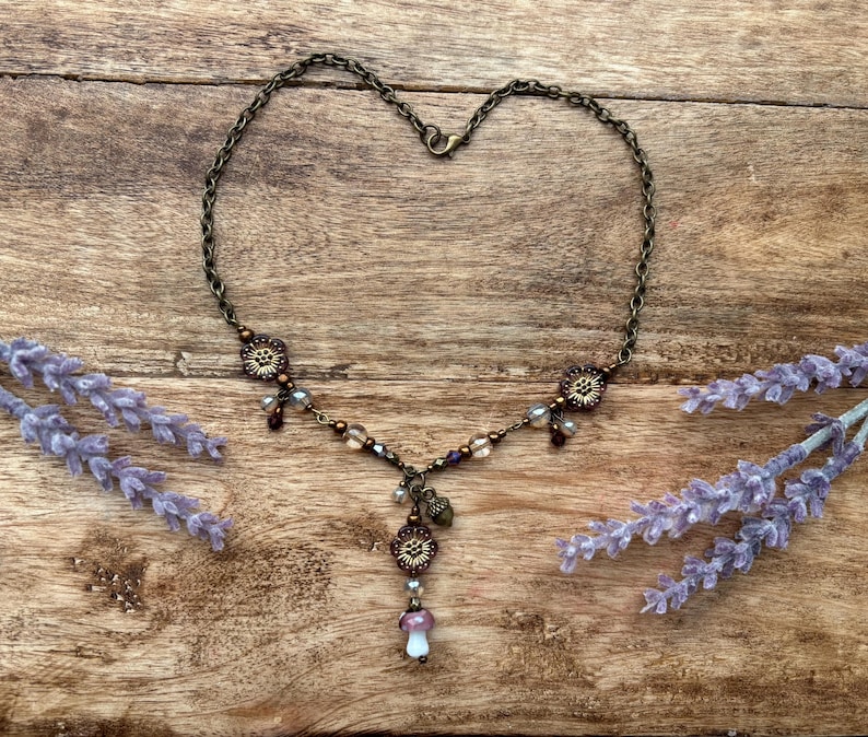 May include: A bronze necklace with a heart-shaped chain. It features floral and mushroom pendants, plus small beads in purple, brown, and clear. Lavender sprigs are placed on a wooden surface.