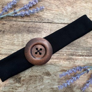 May include: A black elastic headband featuring a large, circular, dark brown wooden button centered on the fabric. The button has a smooth, polished finish with four small holes in the middle arranged in a square pattern for threading.