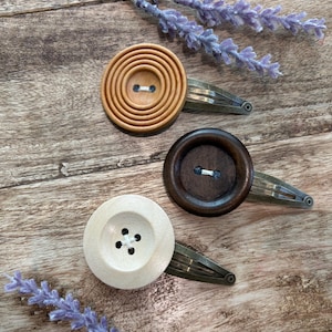 Pode incluir: Três ganchos de cabelo com botões de madeira circulares nas cores castanho-claro, castanho-escuro e creme. Cada botão está preso a um gancho de cabelo metálico de pressão. O botão castanho-claro tem sulcos circulares concêntricos.