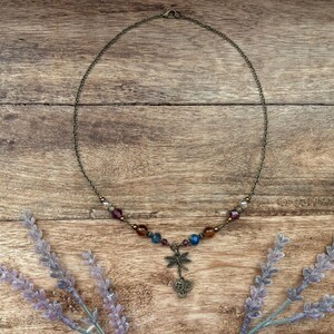 Puede incluir: Un delicado collar con un colgante de lib&eacute;lula y cuentas de colores. El collar presenta una cadena de color bronce y un dije de lib&eacute;lula con detalles intrincados. Las cuentas son en tonos morado, azul y &aacute;mbar. El collar se exhibe sobre una superficie de madera.