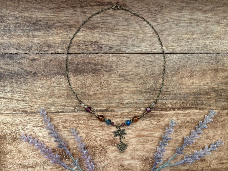 Puede incluir: Un delicado collar con un colgante de lib&eacute;lula y cuentas de colores. El collar presenta una cadena de color bronce y un dije de lib&eacute;lula con detalles intrincados. Las cuentas son en tonos morado, azul y &aacute;mbar. El collar se exhibe sobre una superficie de madera.