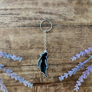 May include: A black silhouette keychain of a figure with a silver chain and ring. The keychain is set against a wooden background with lavender sprigs on either side. The figure is a dark color with a light blue outline.