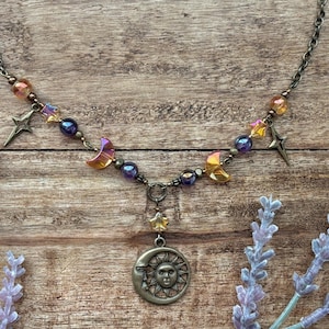 Op de afbeelding: Een ketting met een hemels thema met een bronzen zon- en maan hanger. De ketting bevat ster- en halve maan bedels, samen met iriserende kralen in paars, oranje en geel. De ketting is donker bronskleurig.