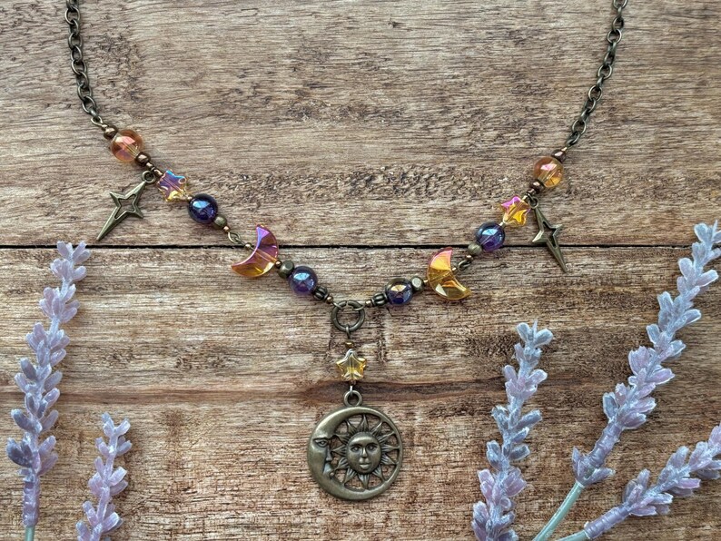 Op de afbeelding: Een ketting met een hemels thema met een bronzen zon- en maan hanger. De ketting bevat ster- en halve maan bedels, samen met iriserende kralen in paars, oranje en geel. De ketting is donker bronskleurig.