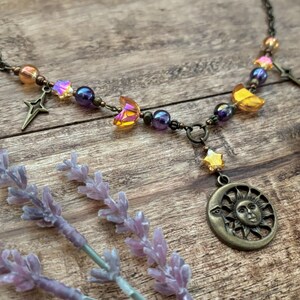 Op de afbeelding: Een ketting met een hemels thema met een bronzen zon- en maanhanger, ster- en maansikkelsieraden en iriserende kralen. De ketting wordt gepresenteerd op een houten oppervlak met lavendeltakjes.