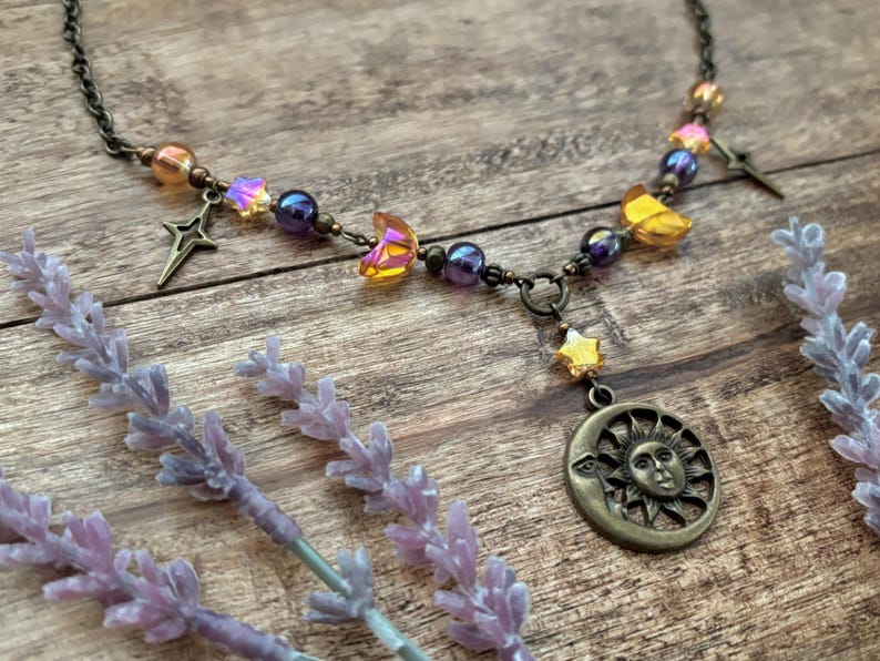 Op de afbeelding: Een ketting met een hemels thema met een bronzen zon- en maanhanger, ster- en maansikkelsieraden en iriserende kralen. De ketting wordt gepresenteerd op een houten oppervlak met lavendeltakjes.