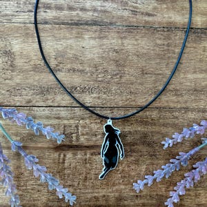 Può includere: Una collana con pendente nero a forma di silhouette femminile. Il pendente è appeso a un cordino nero. Rami di lavanda sono disposti attorno alla collana su una superficie di legno.