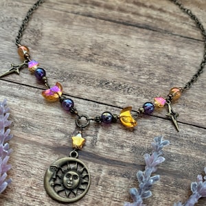 Op de afbeelding: Een ketting met hemelse thema's met een bronzen ketting en hangers. De ketting heeft een zon- en maanhanger, sterbedels en iriserende kralen in paars, oranje en geel. De ketting wordt gepresenteerd op een houten oppervlak.