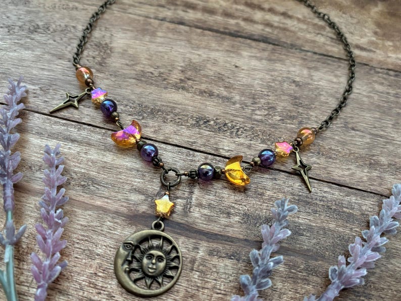 Op de afbeelding: Een ketting met hemelse thema's met een bronzen ketting en hangers. De ketting heeft een zon- en maanhanger, sterbedels en iriserende kralen in paars, oranje en geel. De ketting wordt gepresenteerd op een houten oppervlak.