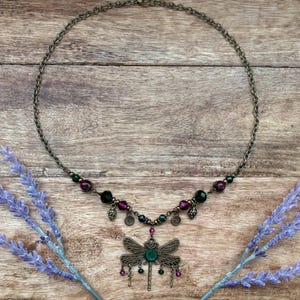 Puede incluir: Un collar de tono bronce con un colgante de libélula, una piedra central verde y detalles colgantes. El collar incluye cuentas moradas y verdes, sobre un fondo de madera con ramitas de lavanda.