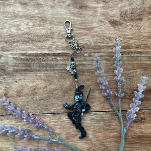 Puede incluir: Un llavero negro y plateado con un mosquetón, un dije de araña, un dije de calavera y una figura de banda de música. El llavero está sobre un fondo de madera con ramitas de lavanda.