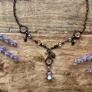 May include: A bronze necklace with floral and bead details, including a mushroom pendant. The necklace is displayed on a wooden surface with lavender. The chain length is approximately 46 cm.