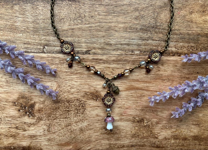 May include: A bronze necklace with floral and bead details, including a mushroom pendant. The necklace is displayed on a wooden surface with lavender. The chain length is approximately 46 cm.