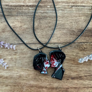 May include: Two black pendant necklaces featuring a couple kissing with red accents. The pendants hang from black cords. The image is set against a wooden background with lavender sprigs.